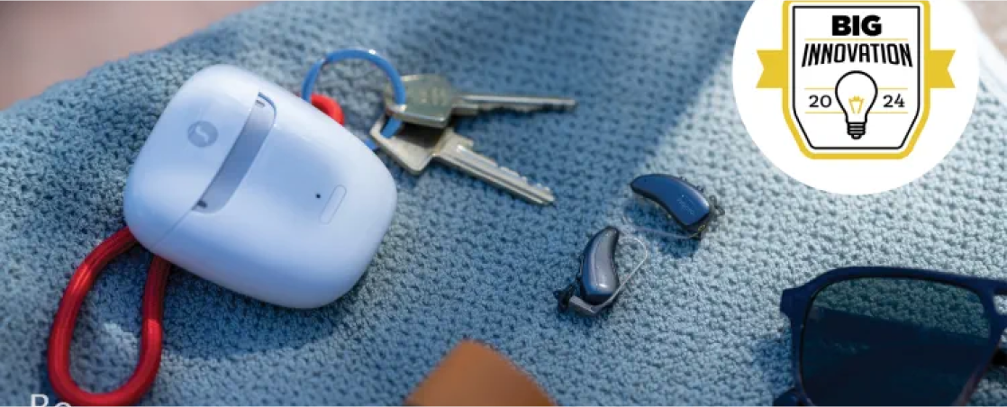 Close-up of a hearing aid set lying on a textured blue surface, alongside a white charging case, a set of keys, and sunglasses. A "Big Innovation 2024" award badge is in the top right corner.