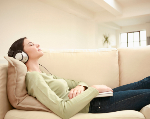 A person lies on a sofa with their head on a cushion, wearing headphones and a relaxed expression. They are holding a book on their lap. The room is softly lit, with a large window and a plant in the background.