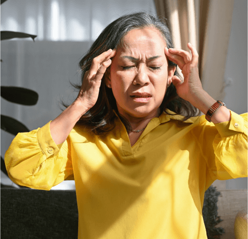 A woman in a yellow blouse sits with her eyes closed, touching her temples with both hands. She appears to be experiencing discomfort or a headache. Sunlight filters through a window, partially illuminating her face.