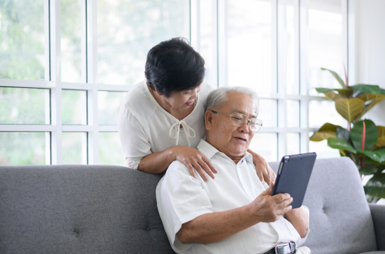 An elderly couple sits on a gray sofa. The man is holding a tablet, and the woman leans over his shoulder, smiling. They are in a bright room with large windows and a plant in the background.