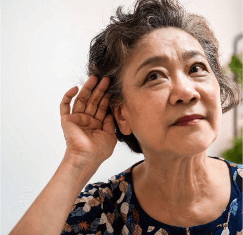 Elderly woman with short gray hair holds her hand to her ear, appearing to listen intently. She wears a floral-patterned top and looks slightly upwards, with a thoughtful expression. The background is softly blurred.