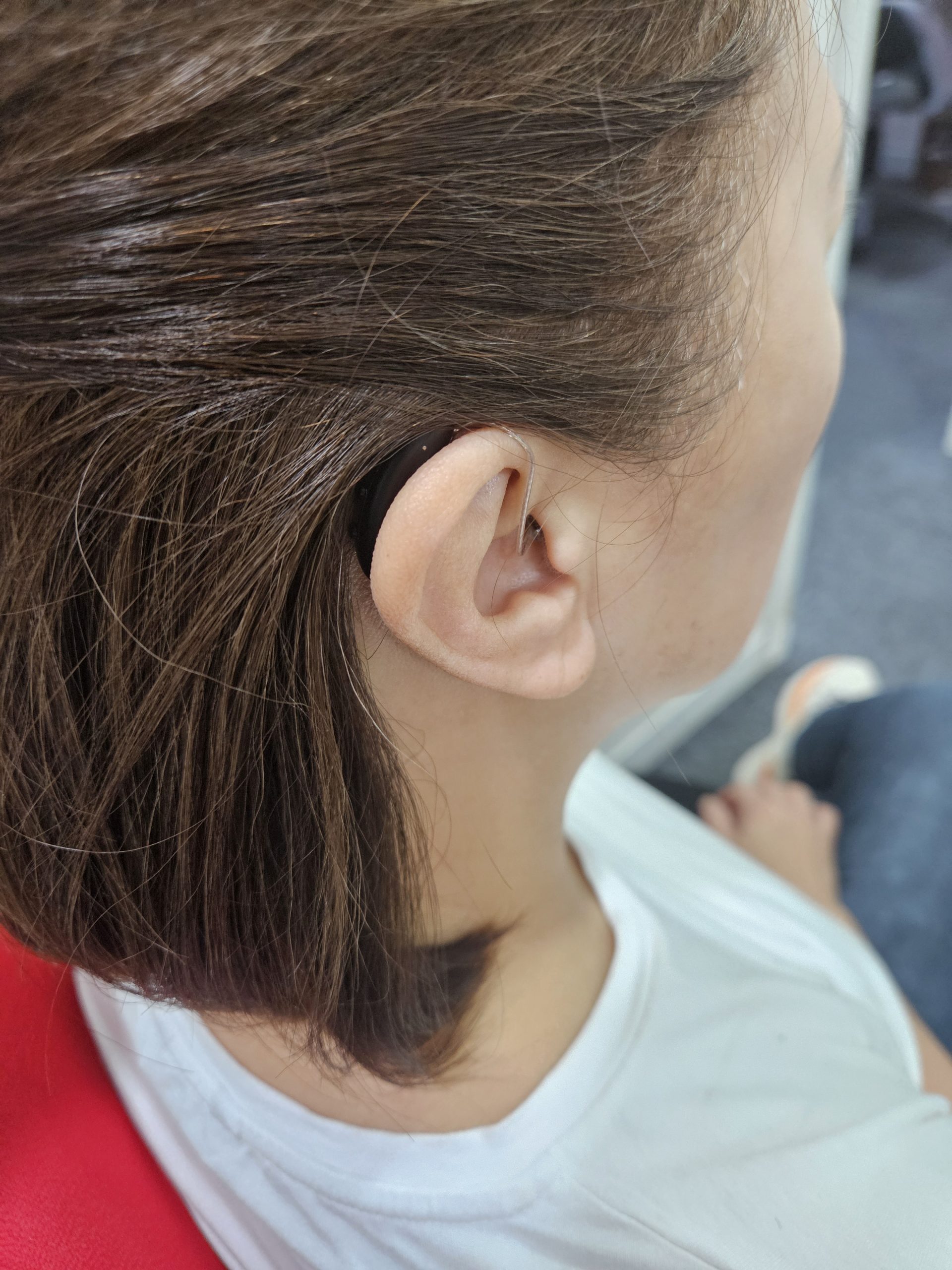 Person with short brown hair wearing a small black behind-the-ear hearing aid, sitting in a chair, wearing a white shirt. The focus is on the hearing aid and the ear.