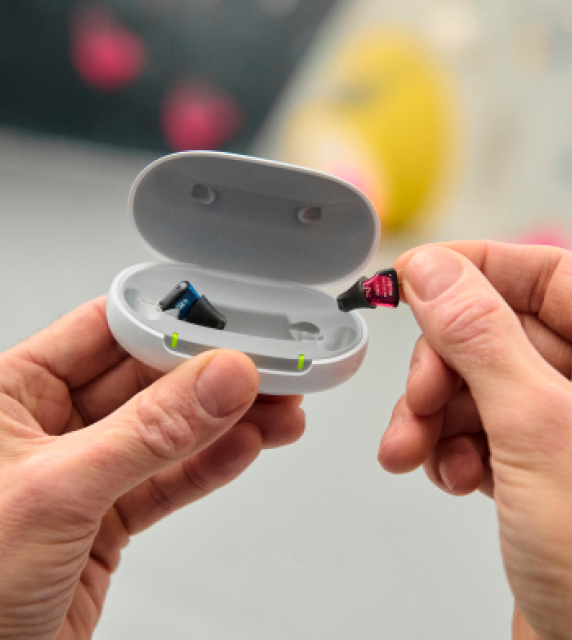 Hands holding a small, open white case containing earbuds. One hand is inserting or removing a small black and red memory card or dongle from the case. The background is blurred with hints of gray and yellow.