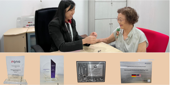 A professional fitting a hearing aid on an elderly woman in an office setting. Below are trophies and plaques, including a glass award and a certificate for professional hearing care.