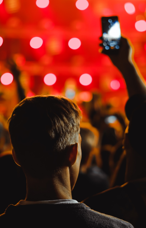 A person with short hair is seen from behind at a concert or event. Their head is silhouetted against bright red stage lights. In the crowd, someone holds up a phone to record the performance.