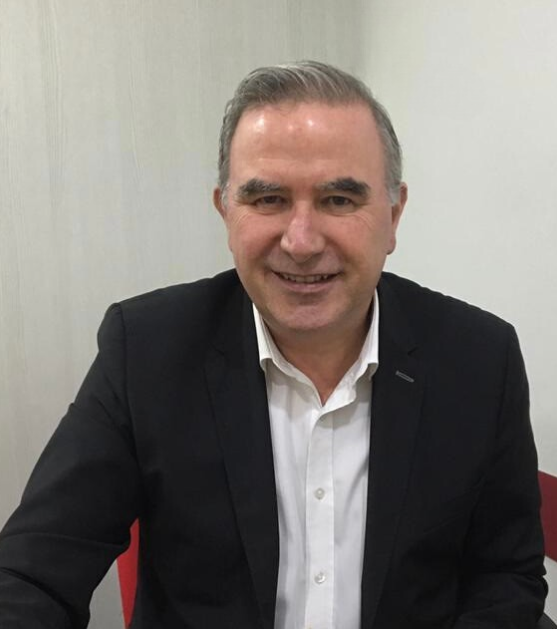 A smiling man with short gray hair is wearing a black blazer over a white shirt. He is seated in a room with light-colored walls.