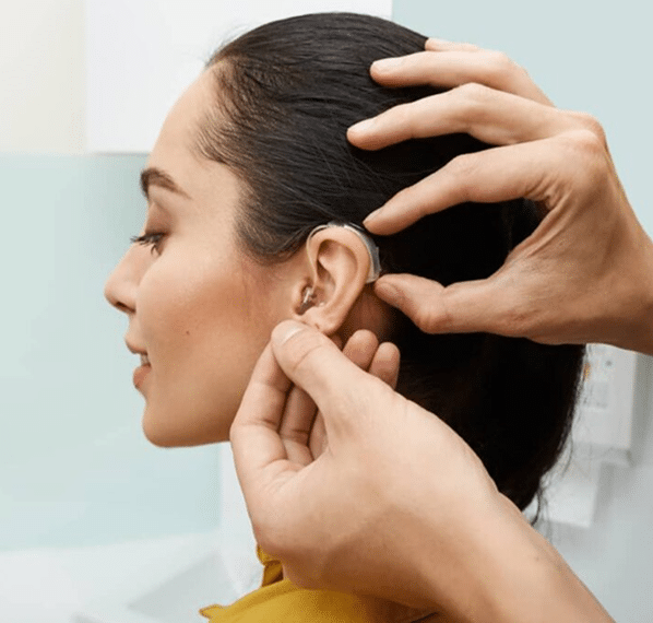 A person fits a behind-the-ear hearing aid onto a woman's ear. The woman is facing left, with her dark hair pulled back, while two hands carefully position the device.