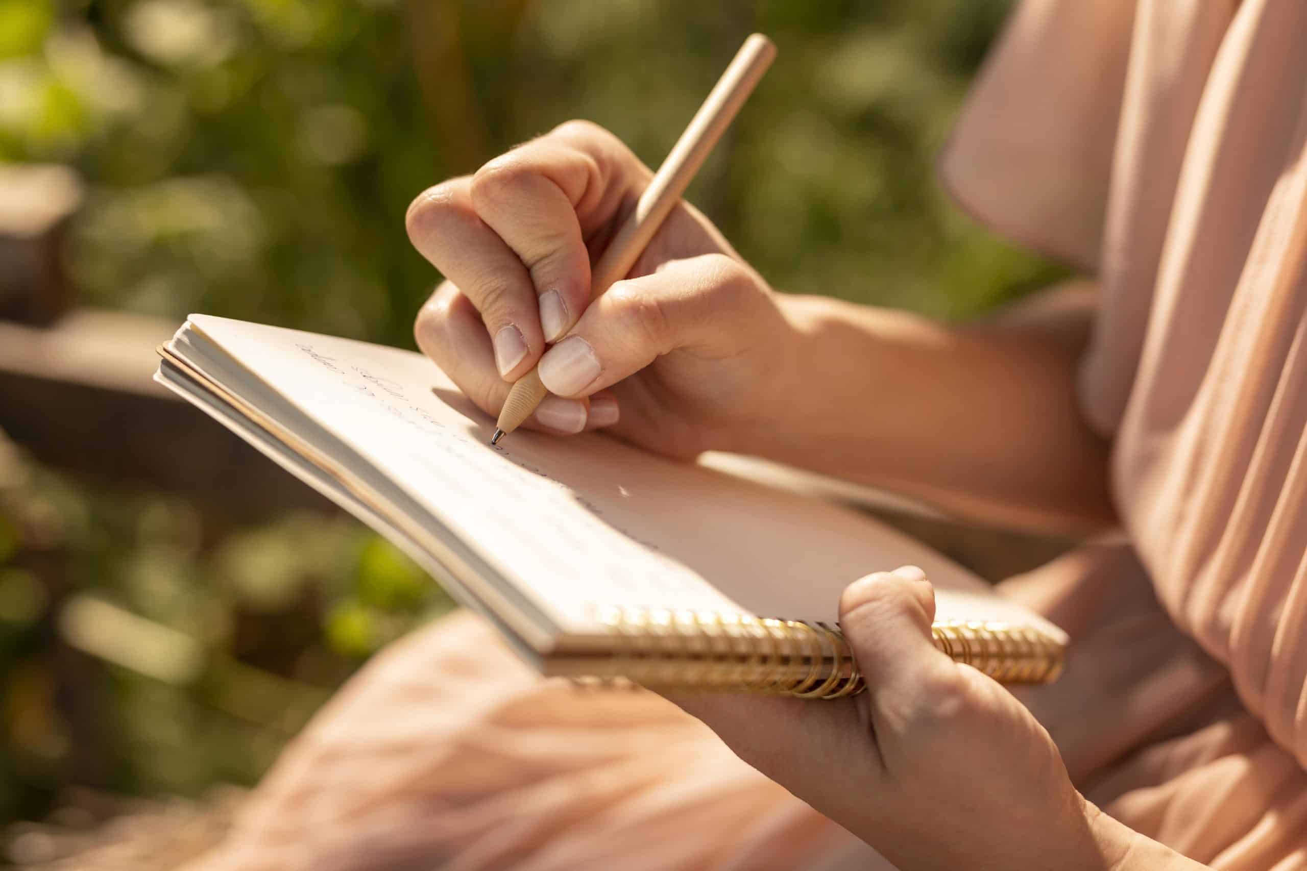 A person holding a spiral notebook and writing with a pencil, seated outdoors in sunlight, with a blurred background of greenery.
