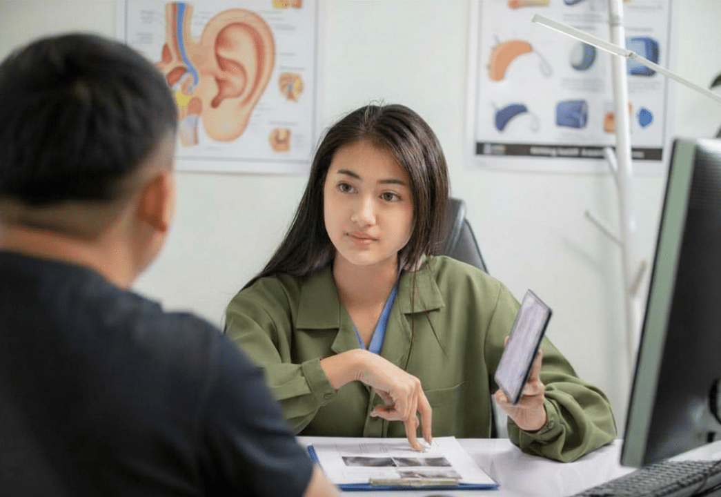 A female doctor in a green coat sits at a desk, holding a tablet and discussing medical images with a patient. Ear anatomy posters are visible on the wall in the background.