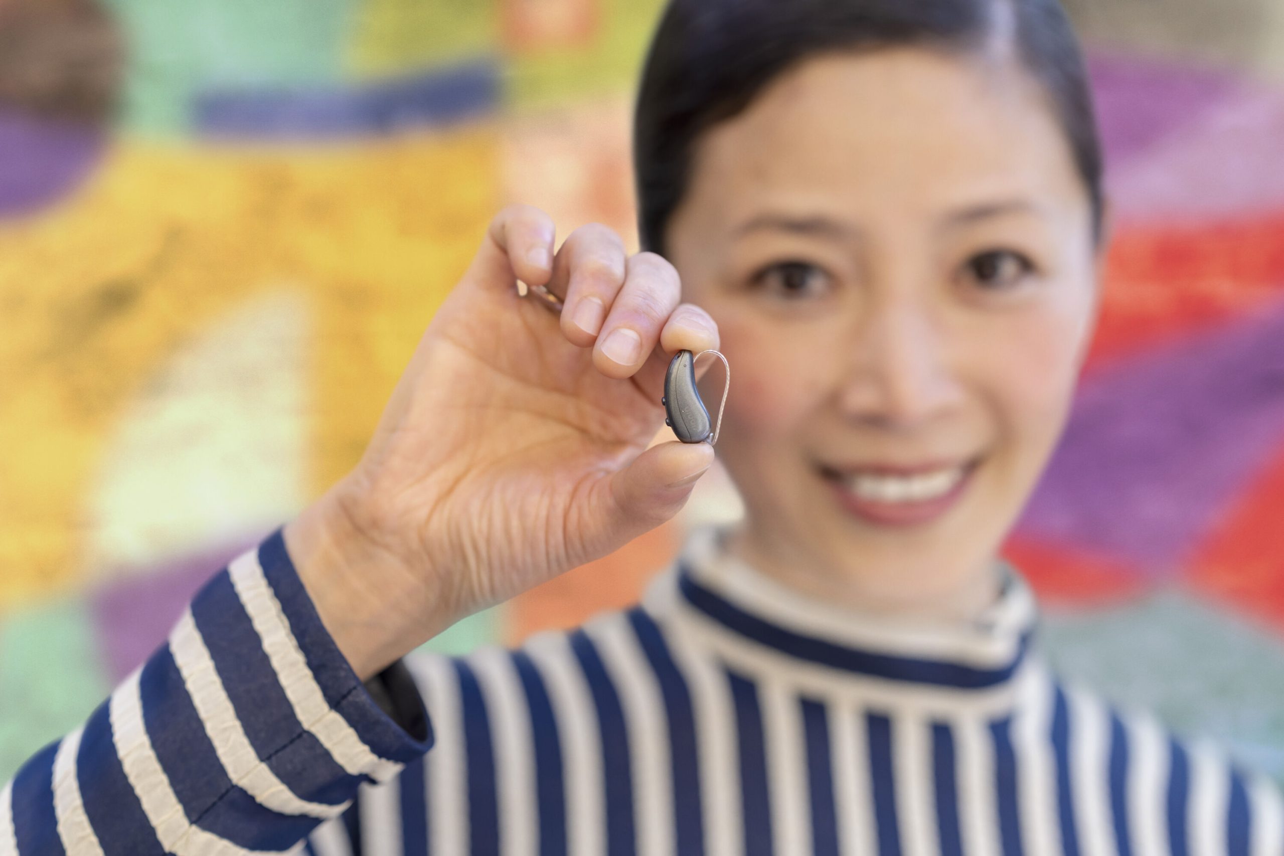 A person in a striped shirt holds a small, hearing aid toward the camera, smiling, with a colorful blurred background.
