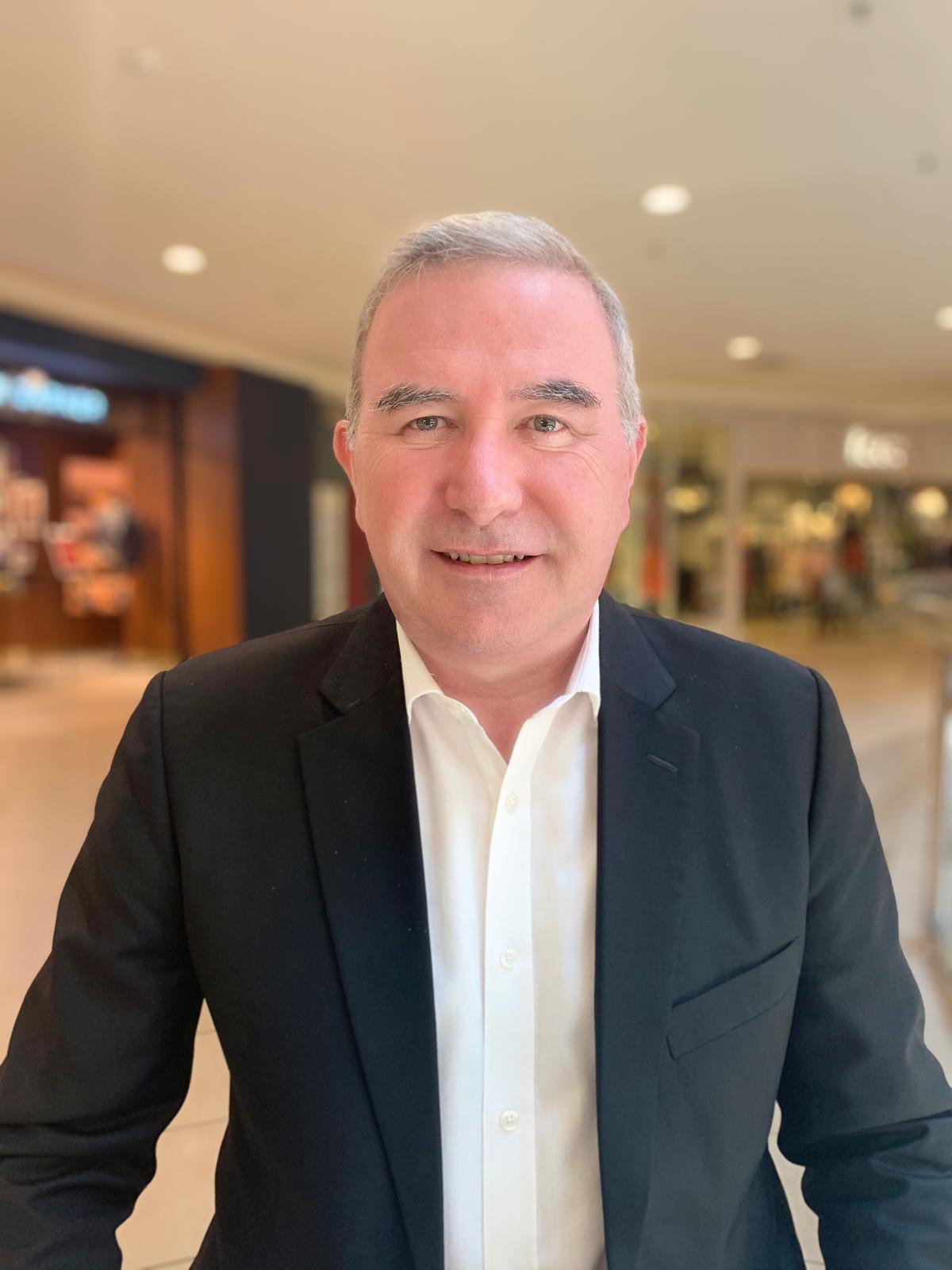 A man with short gray hair wearing a black suit jacket and white shirt stands indoors in a shopping mall, smiling at the camera. The background shows storefronts and blurred lights.