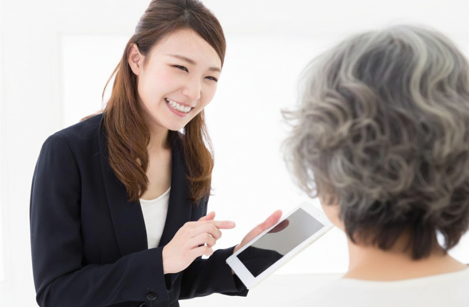 A young woman in a suit smiles and shows a tablet to an older person with gray hair, who is facing away from the camera. The setting is bright and professional.