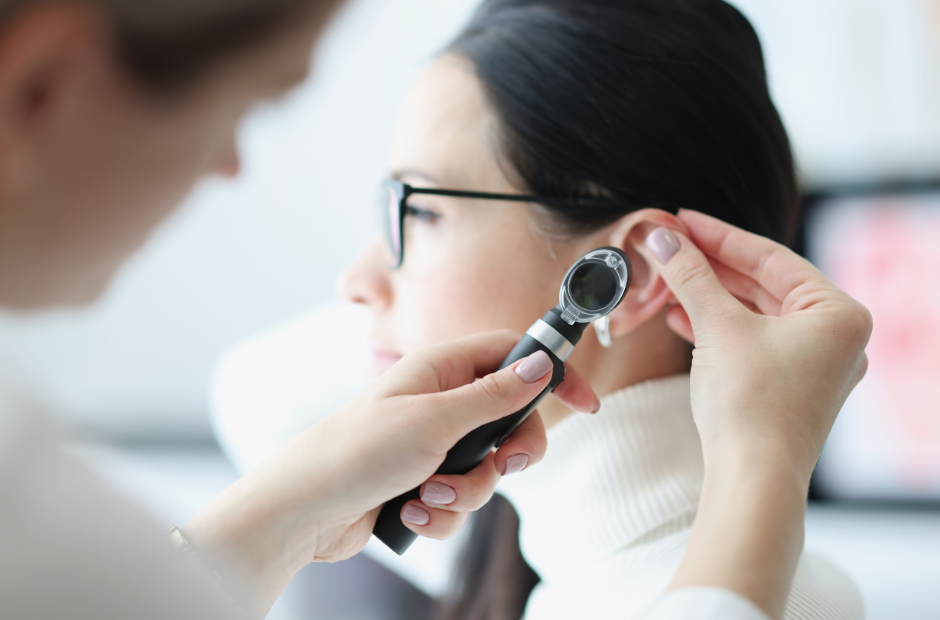 A healthcare professional examines a woman’s ear with an otoscope. The woman wears glasses and a white turtleneck sweater. The background is blurred, focusing on the ear examination.