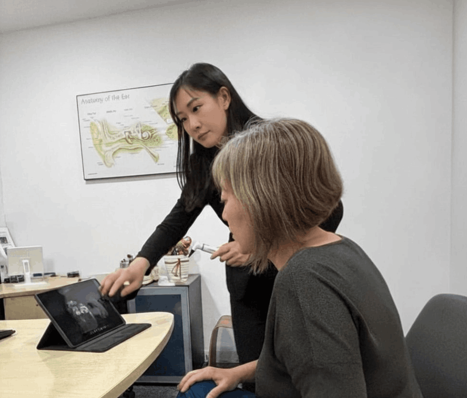 Two women sit at a table. One, seated, looks at a tablet while the other, standing, points at the screen. A diagram labeled “Anatomy of the Ear” is on the wall behind them.