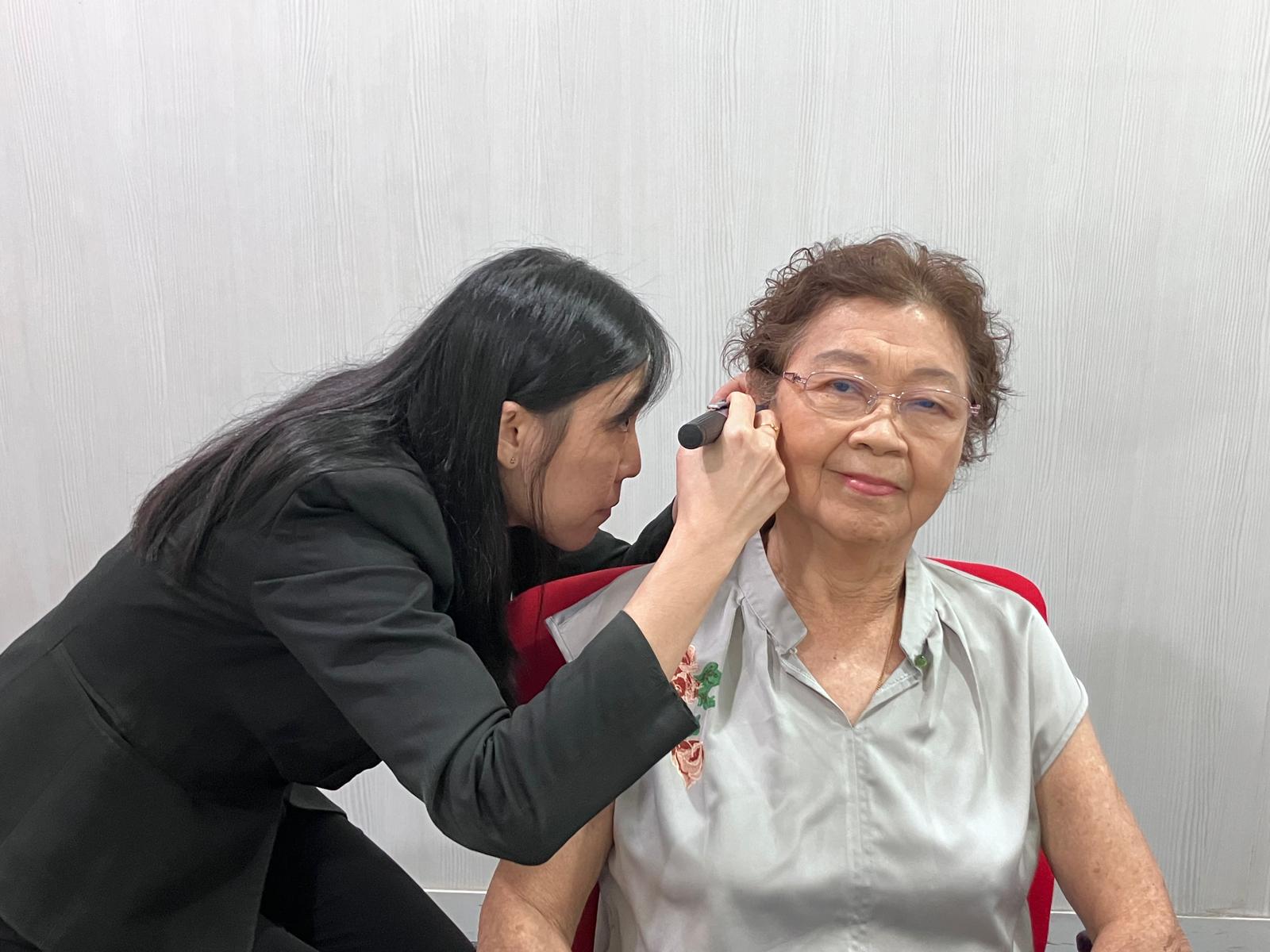 A woman in black examines the ear of an older woman with a medical instrument. The older woman sits on a red chair, looking forward, wearing glasses and a light grey blouse.