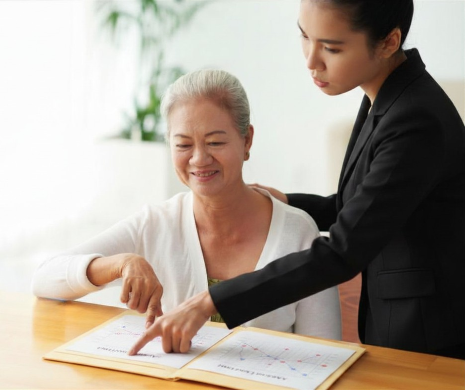 An older woman and a younger woman review documents with graphs together at a table. The younger woman, standing, points at the papers while the older woman, seated, smiles and gestures at the chart.