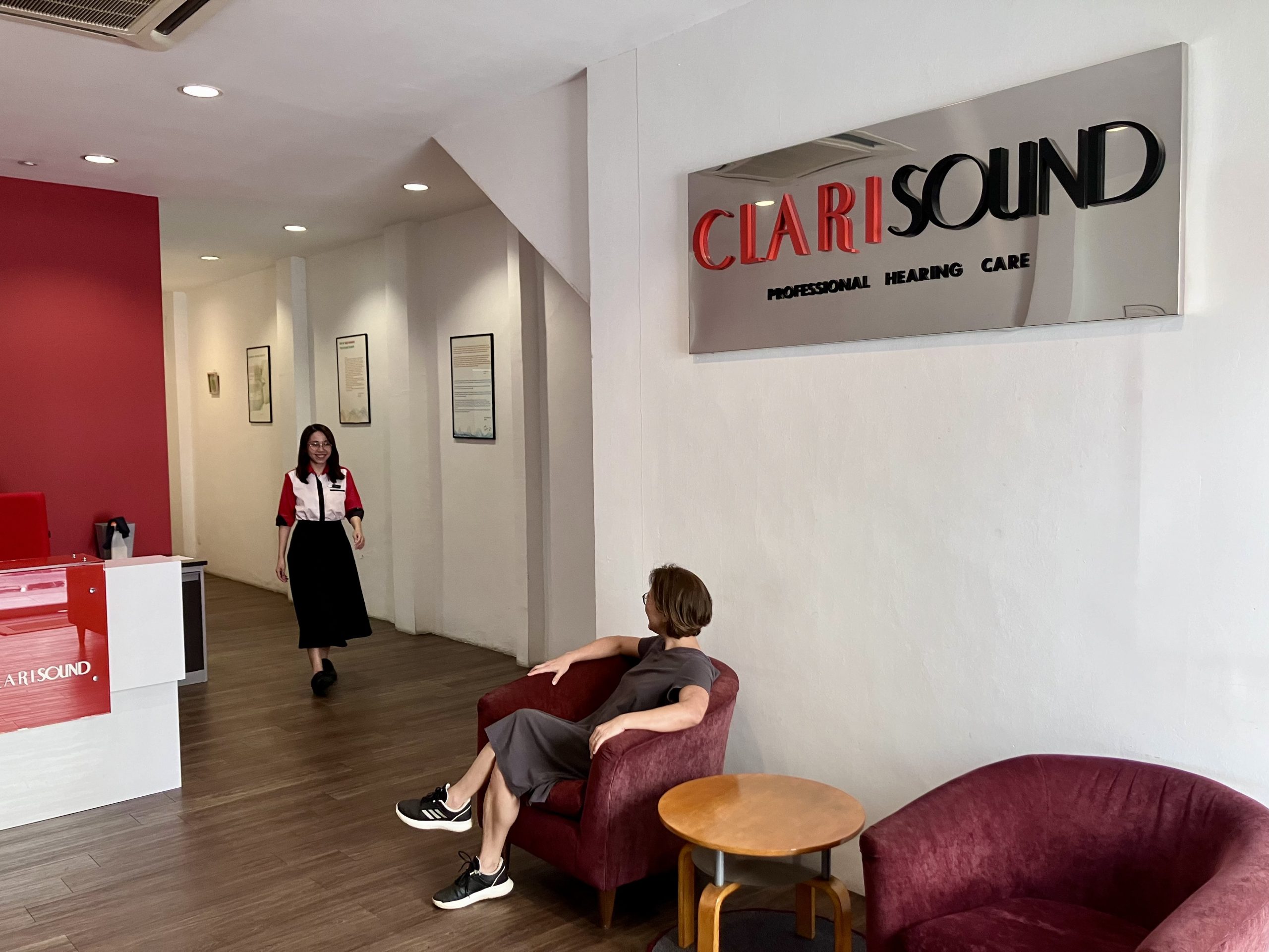 A woman sits on a maroon chair in a waiting area while another woman walks toward her. A sign on the wall reads “CLARISOUND Professional Hearing Care.” The room is modern with white walls and wood floors.