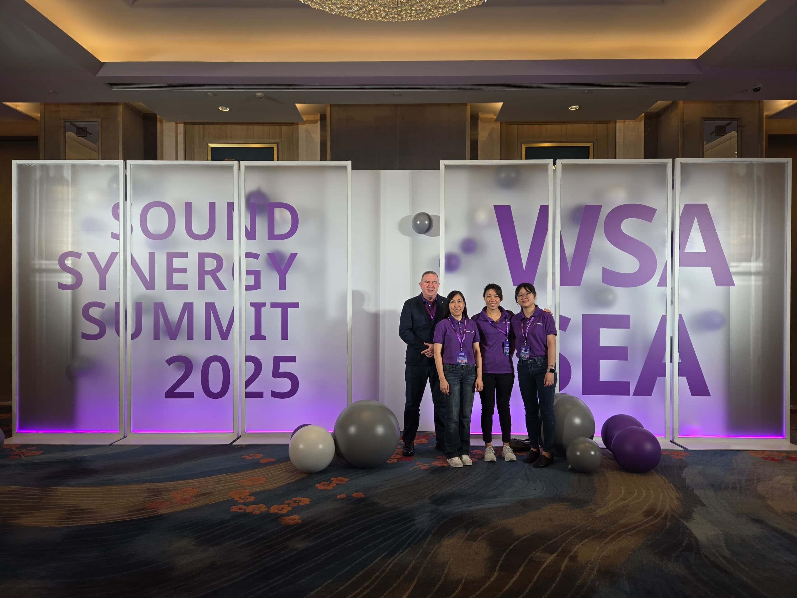 Four people in purple shirts stand smiling in front of a lit backdrop reading “Sound Synergy Summit 2025 WSA SEA,” with several grey and purple balloons on the floor around them.